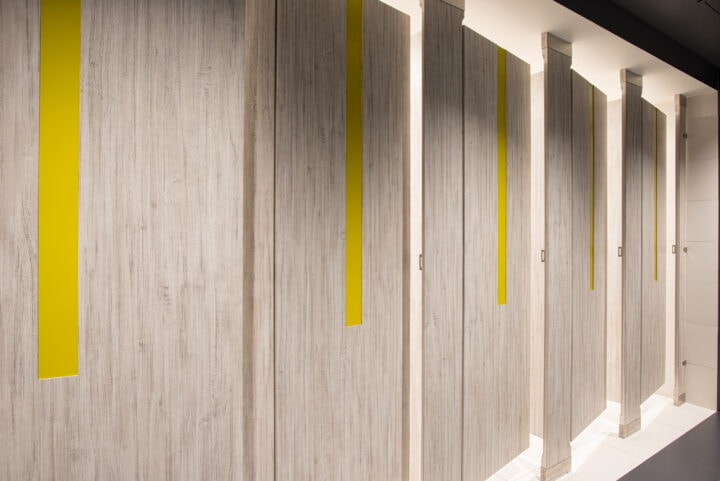 A commercial upscale restroom featuring partitions with high pressure laminate woodgrain doors and pilasters. Floor to ceiling mounted with yellow acrylic inlays in the doors.