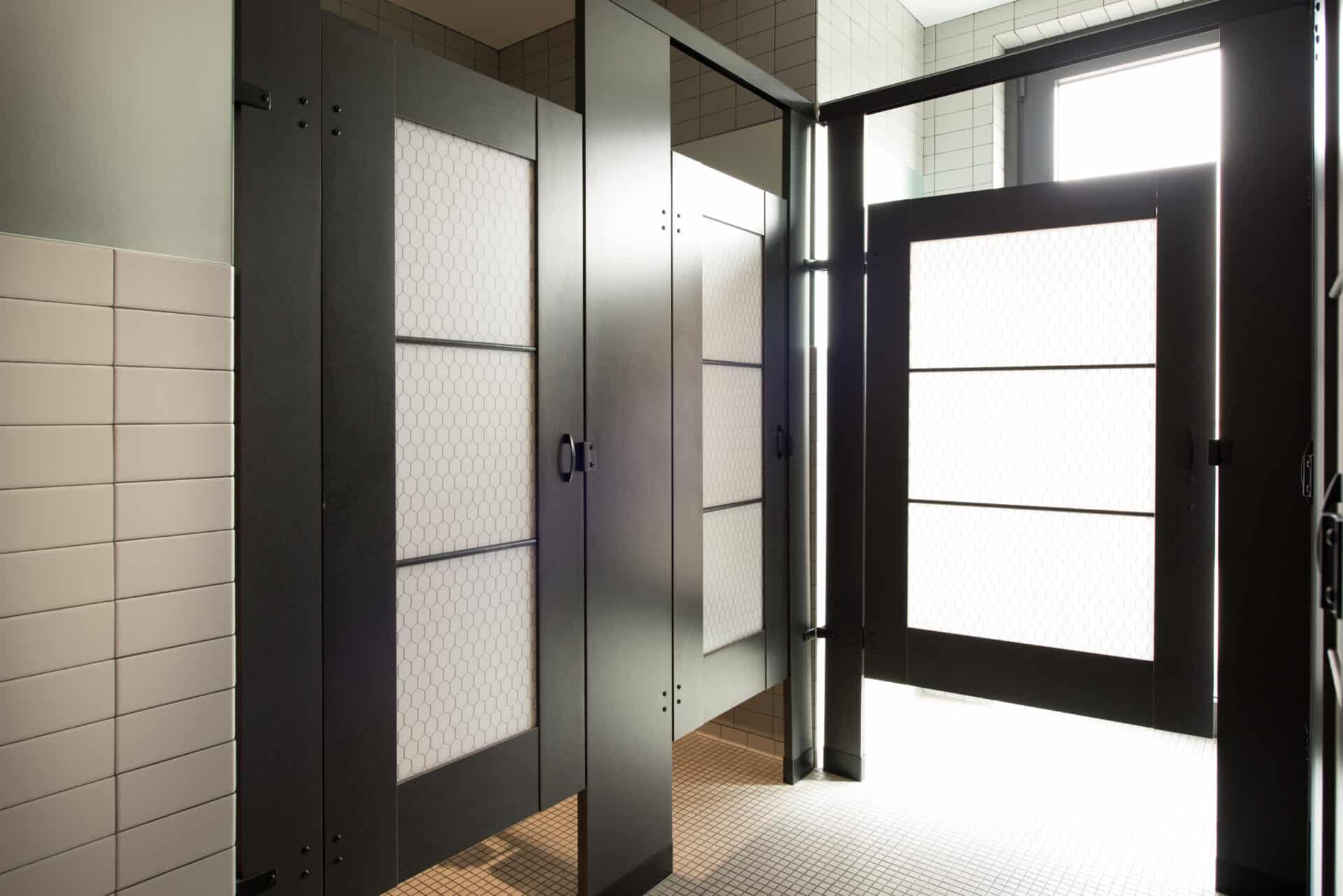 A restroom with three stalls made of high pressure laminate, mounted with powder coated headrail bracing and with chicken wire pattern acrylic door lites.