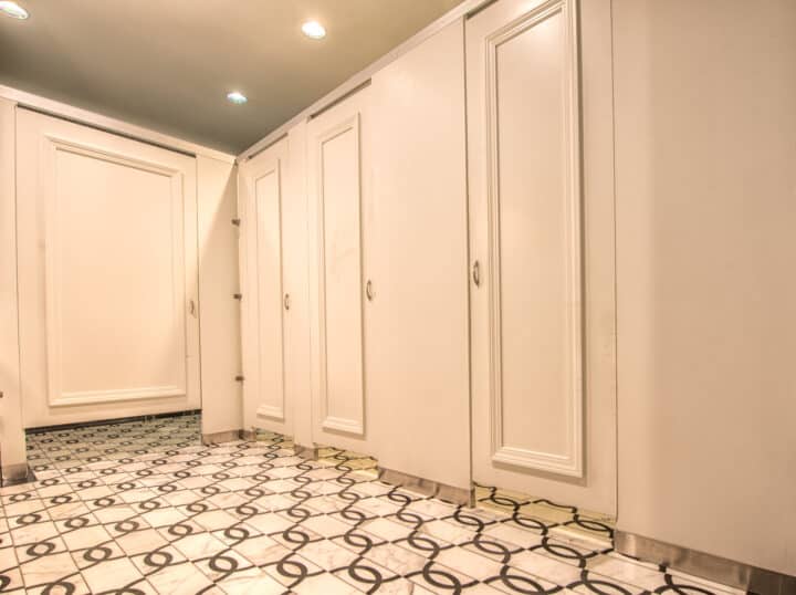 A brightly lit room with several closed white-panel doors, ornate molding lining the walls, and a patterned tile floor.