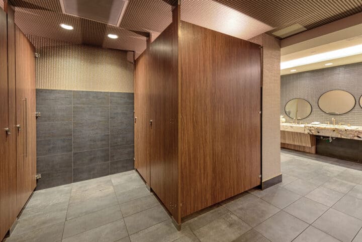 Woodgrain high-pressure laminate partitions in a commercial restroom with modern vanity featuring round mirrors and stone countertops.