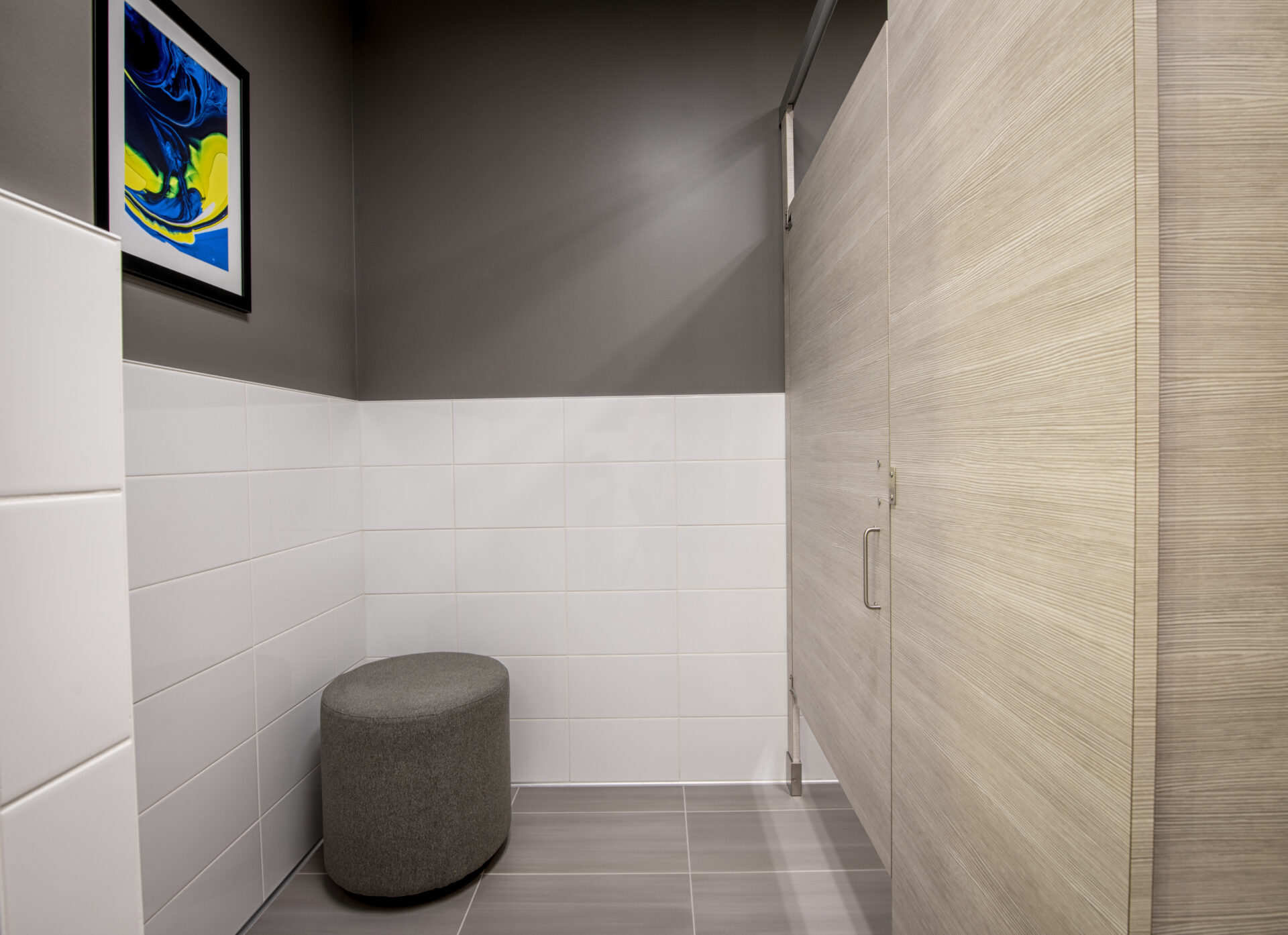 A modern restroom with white tiles, a headrail braced high pressure laminate stall, a gray ottoman, and abstract artwork on the wall.