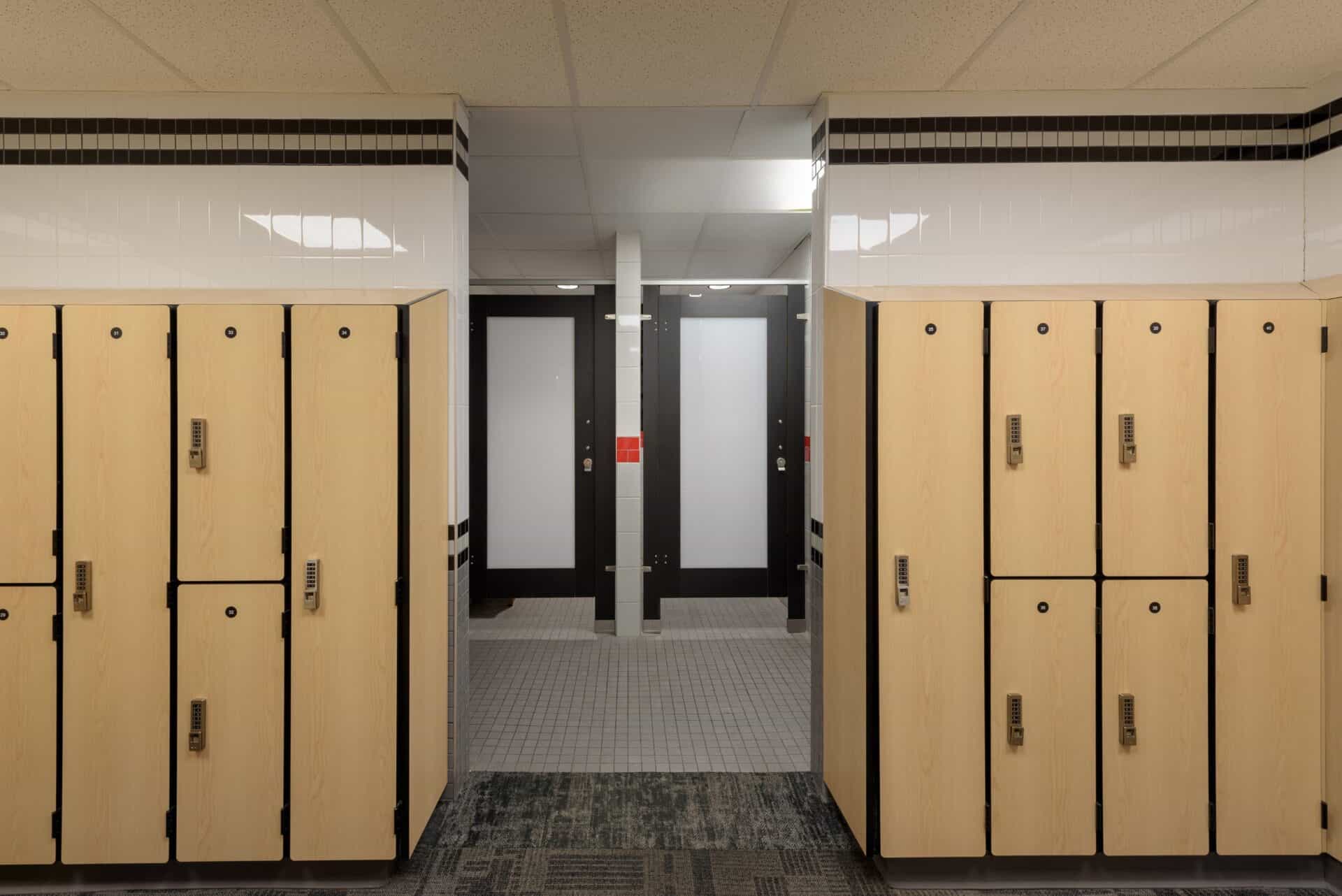 Wooden lockers line a hallway leading to frosted glass doors in a locker room. The ceiling has recessed lighting and the floor is carpeted with a patterned design.