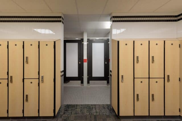 Wooden lockers line a hallway leading to frosted glass doors in a locker room. The ceiling has recessed lighting and the floor is carpeted with a patterned design.