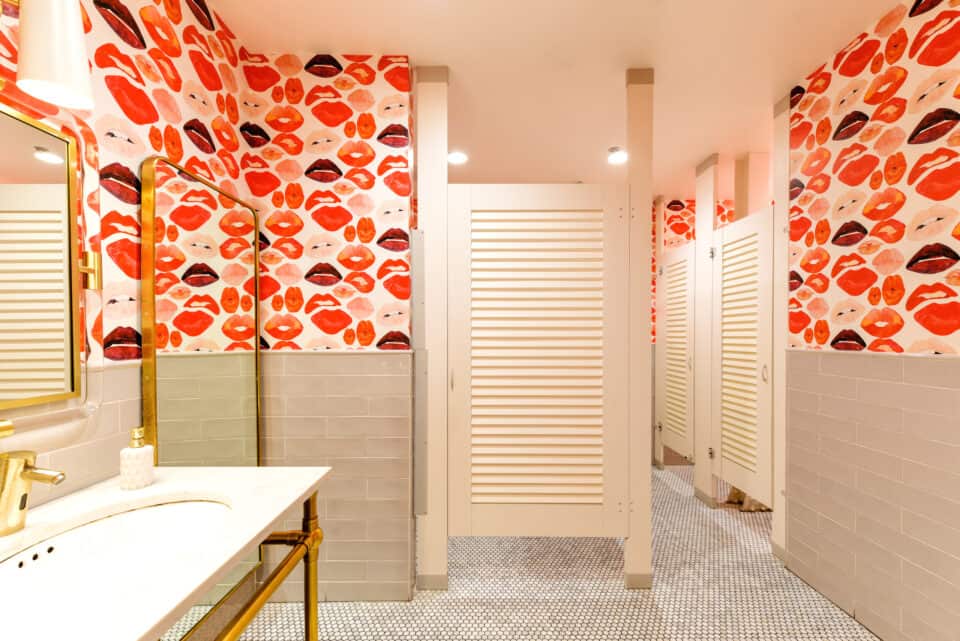 A public restroom with high pressure laminate louver doors mounted floor to ceiling. Bright lips on the wallpaper and sink and mirrors to the left.