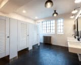 A public restroom with white high pressure laminate louver doors, occupancy indicator latches, floor mounted. Windows and sinks.
