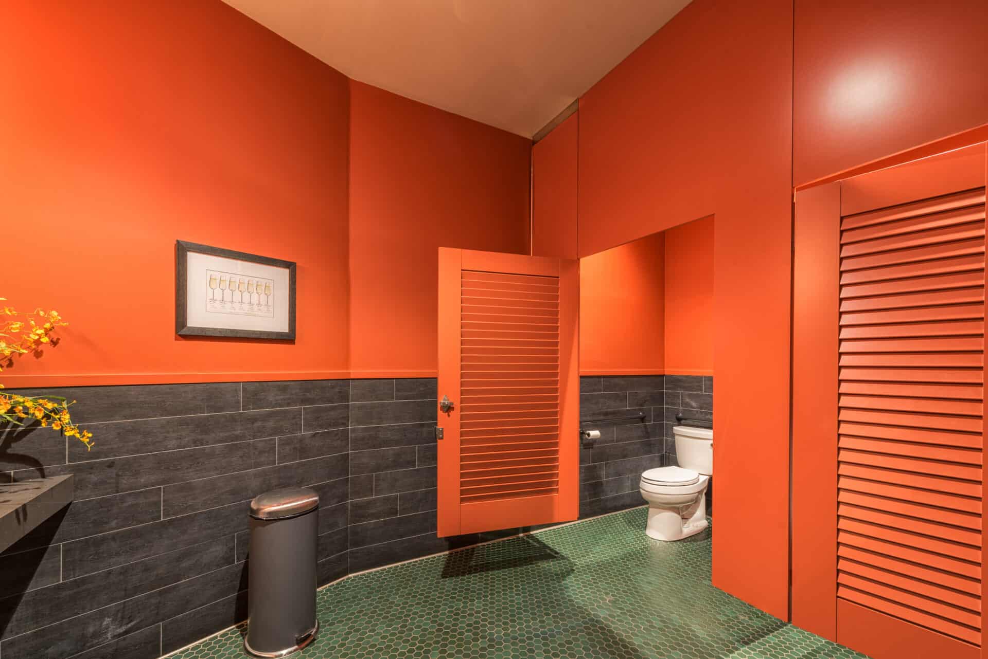 A modern bathroom with orange walls, high pressure laminate painted orange, louvered doors, ceiling hung, gray tile accents, and a green tiled floor. Framed artwork is visible.