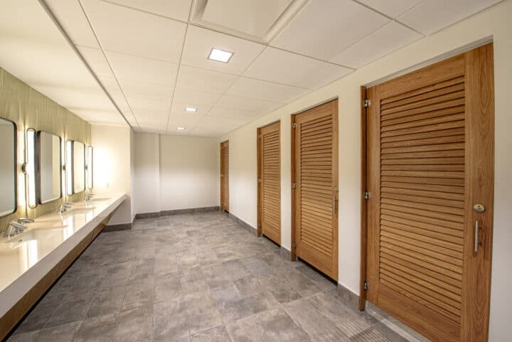Modern commercial restroom with louver doors made from compact laminate, featuring large sinks and polished stone tiles.