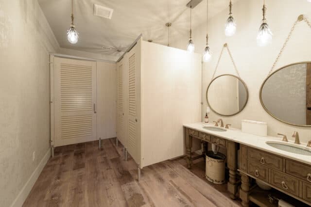 A picture of white high pressure laminate restroom partitions with louvered doors, headrail braced with floor pedestals. Oval mirrors on the right wall and plank wood flooring.
