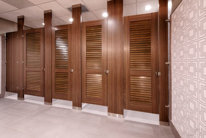 Oversized woodgrain laminate toilet partitions with louvered doors mounted floor to ceiling.