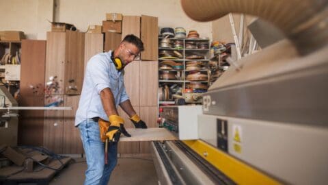A man wearing safety glasses, headphones, and gloves operates a wood-cutting machine in a workshop filled with cabinets and tools, skillfully applying edge banding techniques.