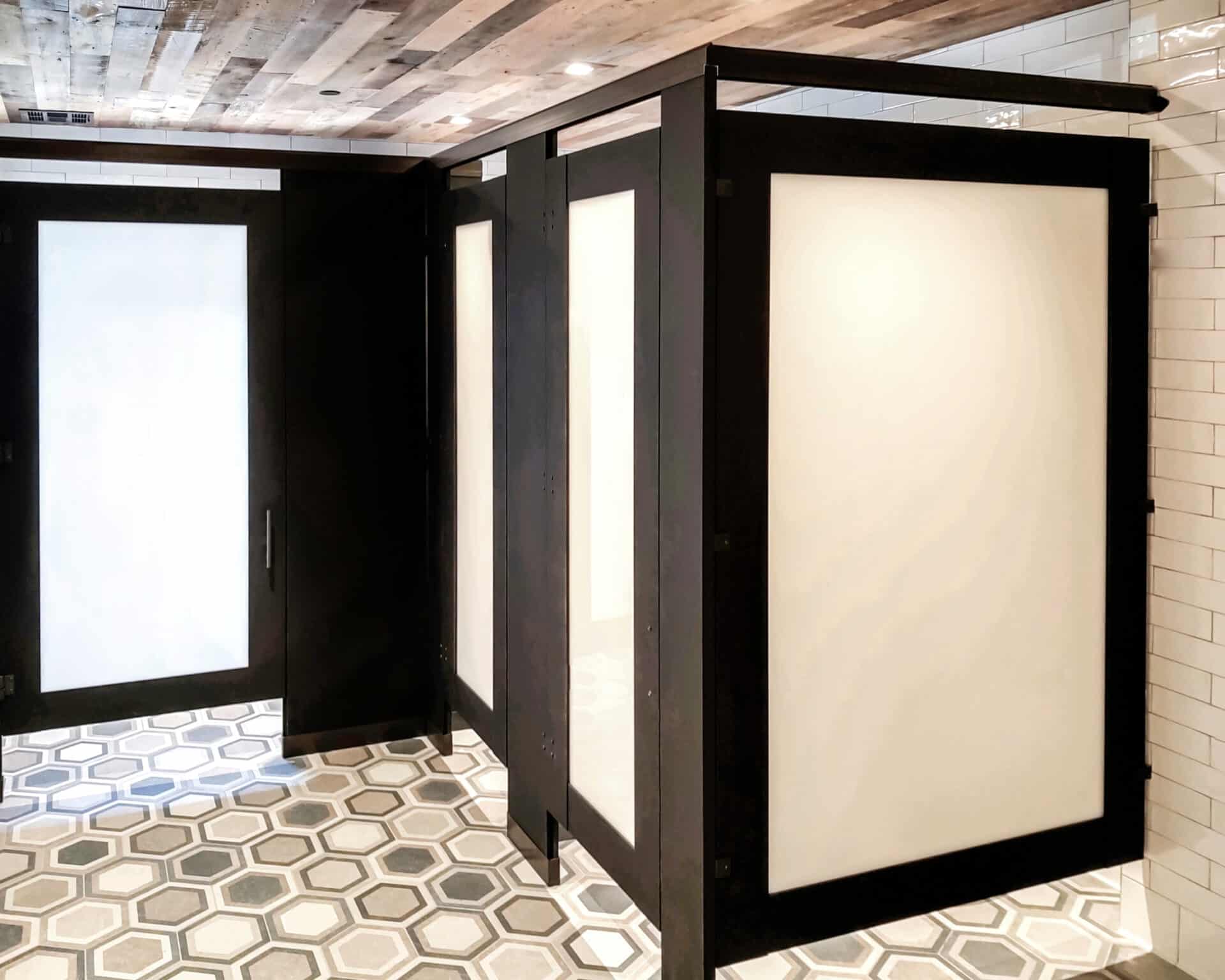 Upscale restroom design with black-framed frosted partitions, stylish hexagon tile flooring, and a reclaimed wood ceiling for a modern industrial look.