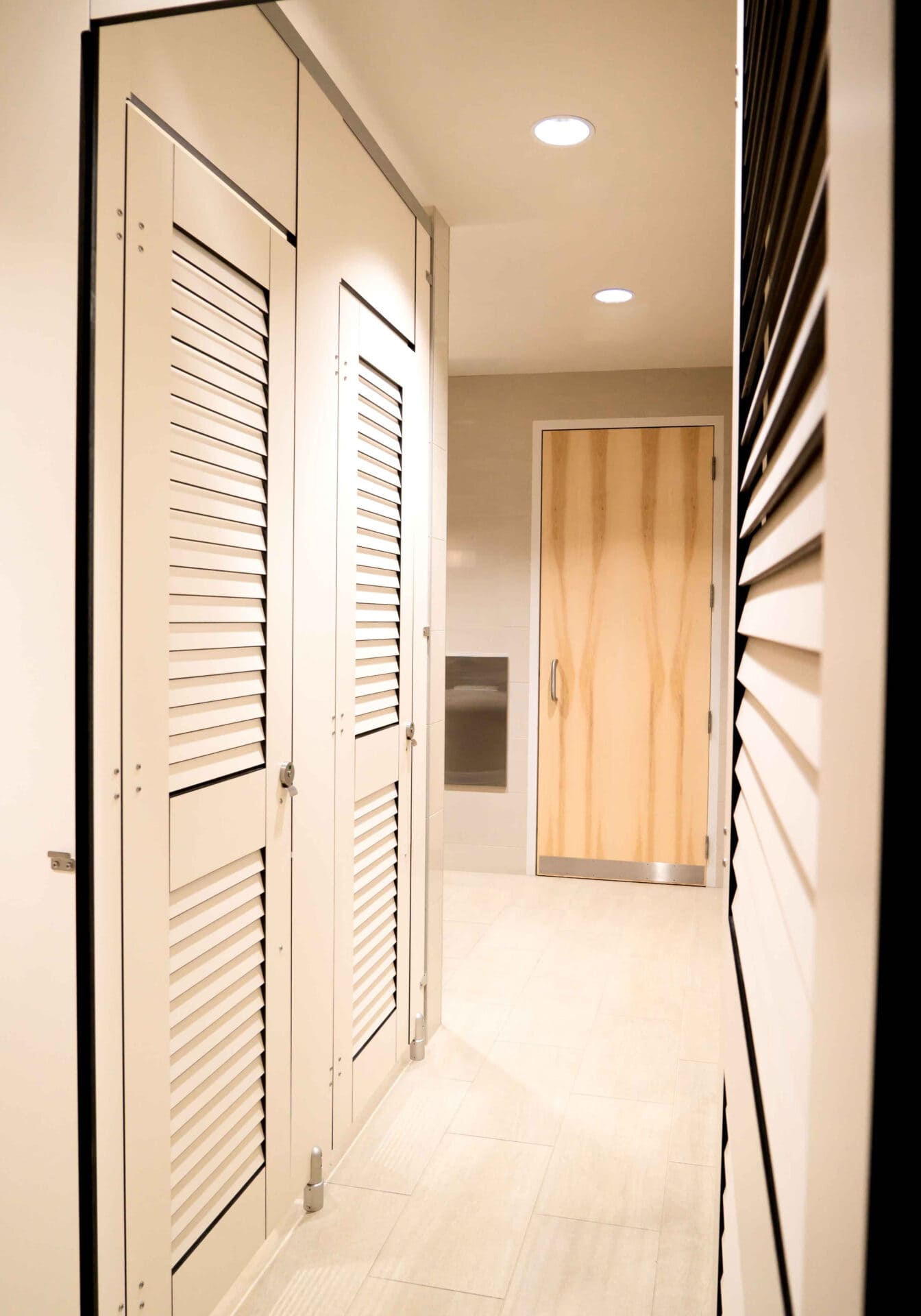 Interior hallway with white louvered doors on the left and a light wood door at the end. The space is well-lit with recessed ceiling lights.