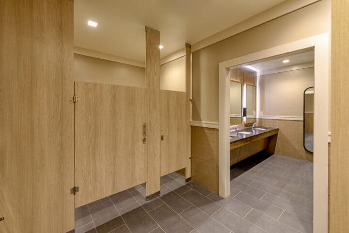 Restroom with compact laminate partitions and floor-to-ceiling pilasters, featuring a modern sink area and tiled flooring.