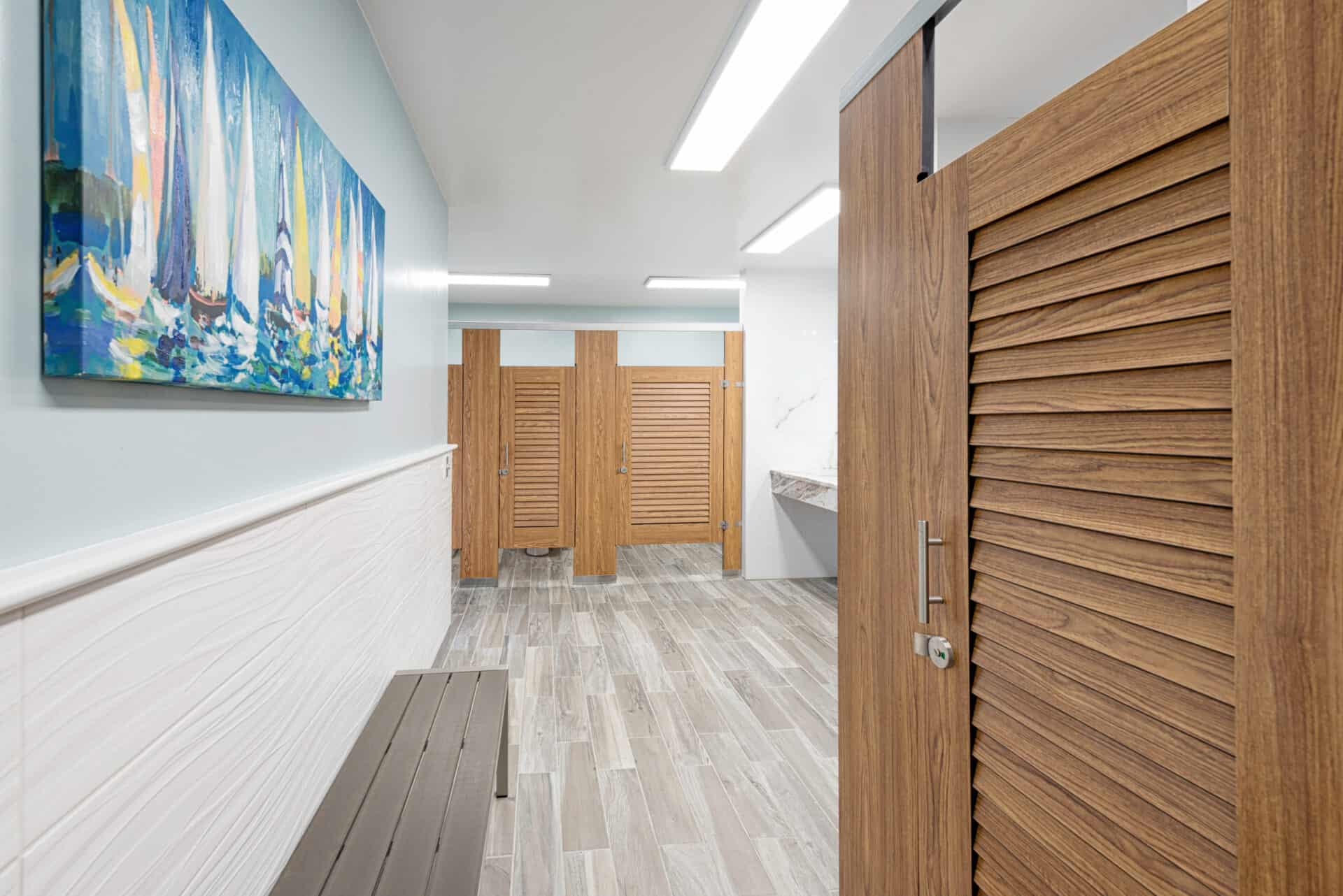 Public restroom with wooden stall doors, a marble countertop with two sinks, and a large mirror above. Bright overhead lighting and tiled floor visible.