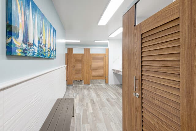 Public restroom with wooden stall doors, a marble countertop with two sinks, and a large mirror above. Bright overhead lighting and tiled floor visible.