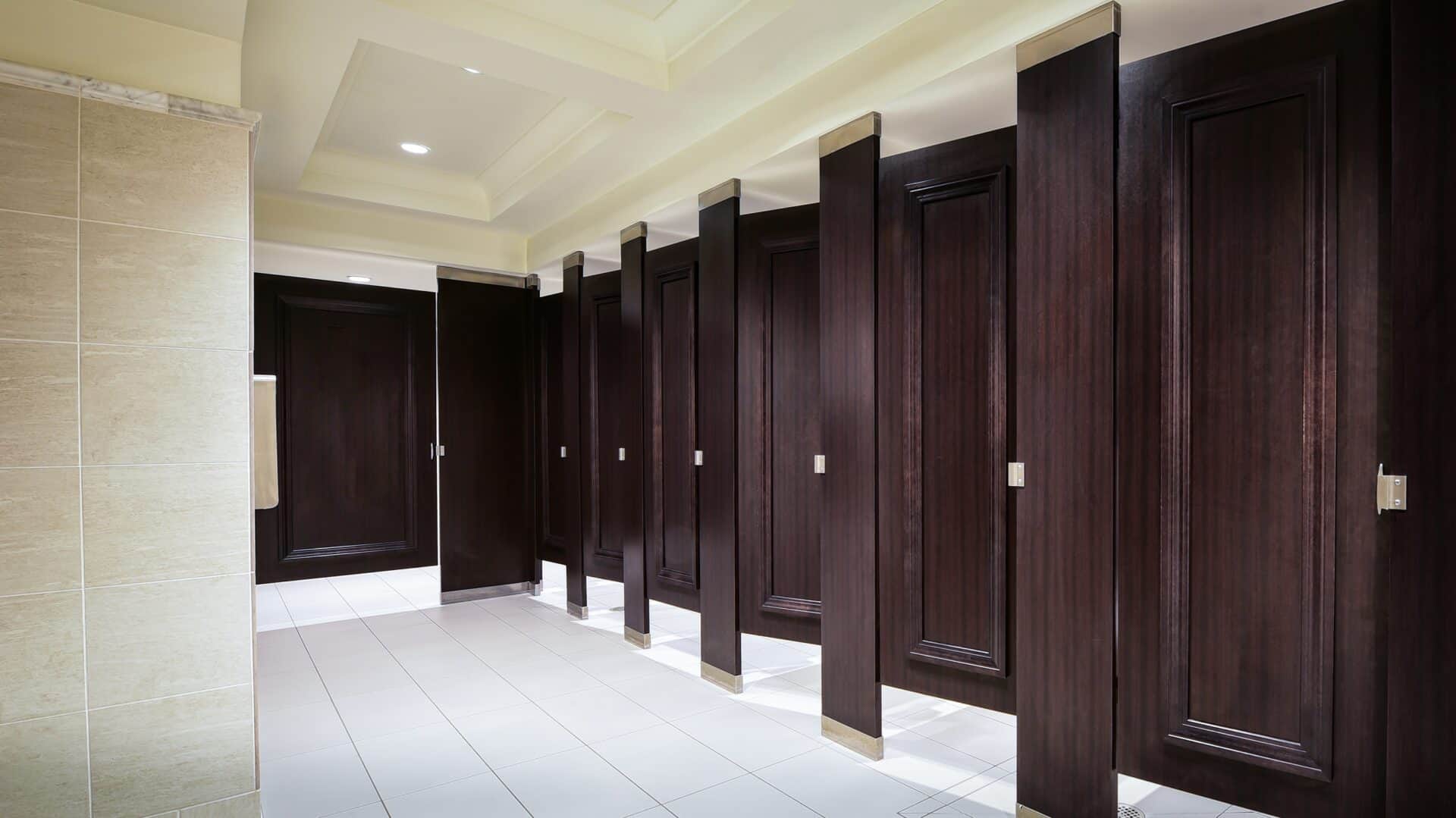 A commercial restroom with a row of partitions in woodgrain plastic laminate, floor to ceiling mounted, with applied moldings on the doors.