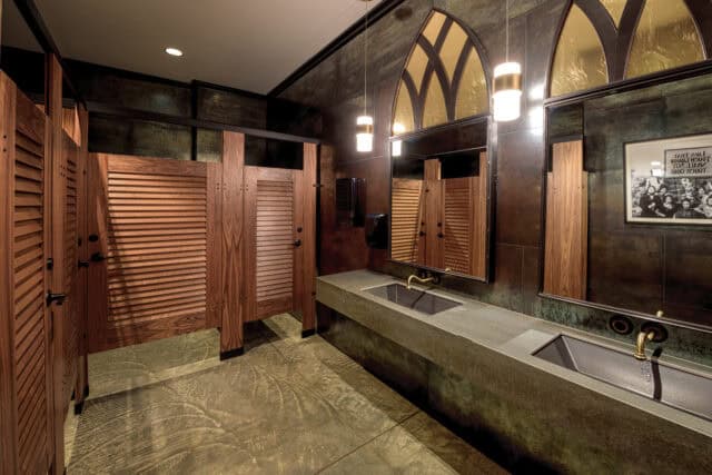 A modern restroom with wooden stall doors, powder coated hardware, a long stone sink equipped with two faucets, mirrored walls, and two wall-mounted lights.