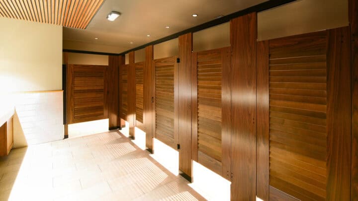 A row of wooden restroom stalls with powder coated headrail braced louver doors stands in a brightly lit bathroom, featuring light-colored tile flooring and a wooden ceiling panel.