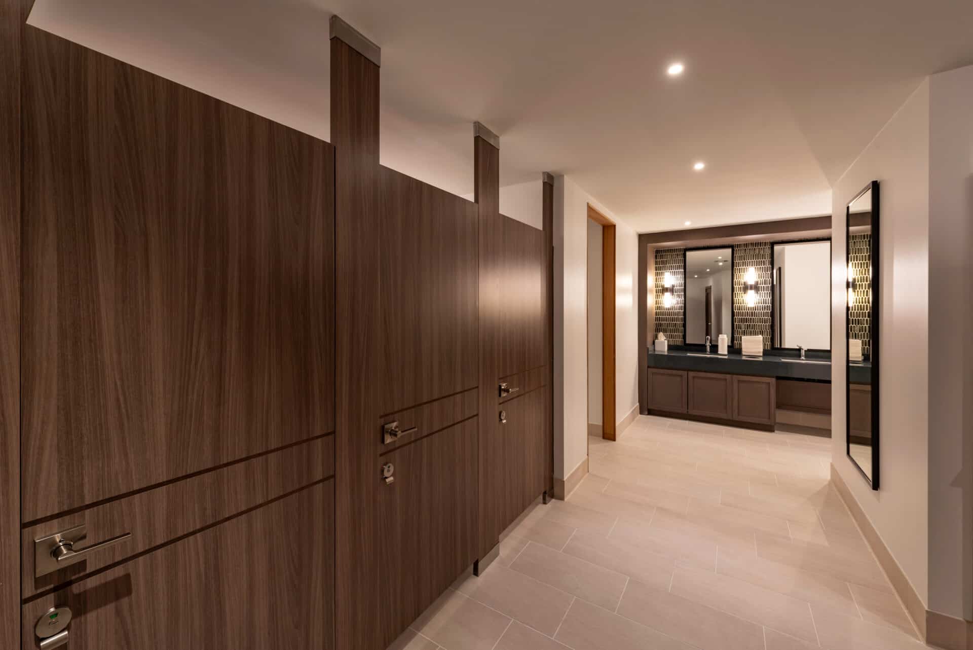 Modern public restroom with wood-paneled stalls on the left and a vanity area with sinks and large mirrors in the background.