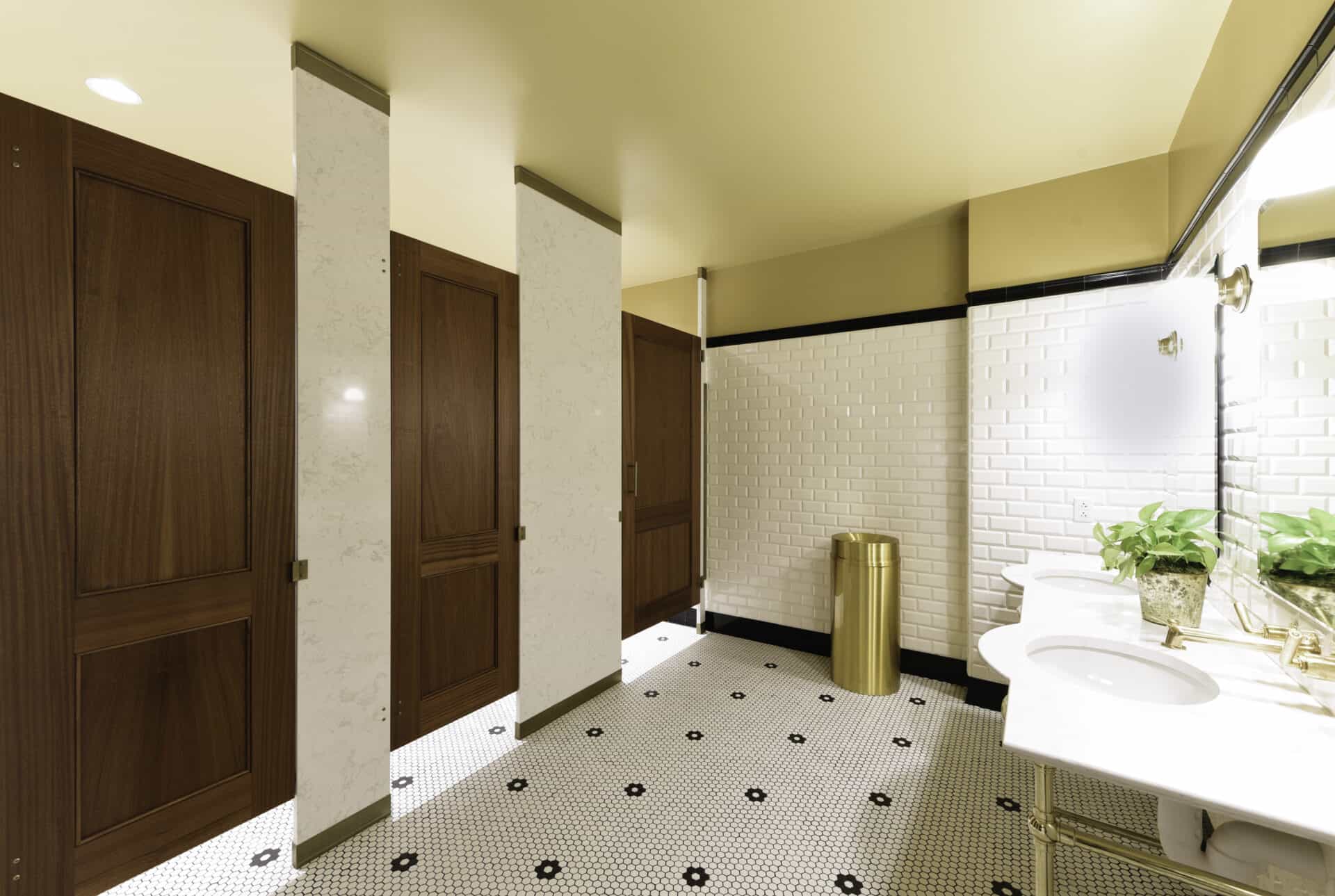 Bathroom with wooden stall doors, white tiled walls, patterned flooring, a countertop with a sink and a potted plant, gold trash can, and a well-lit mirror.