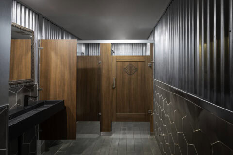 Wood veneer restroom partitions with an engraved door in a rustic industrial bathroom with corrugated metal walls.
