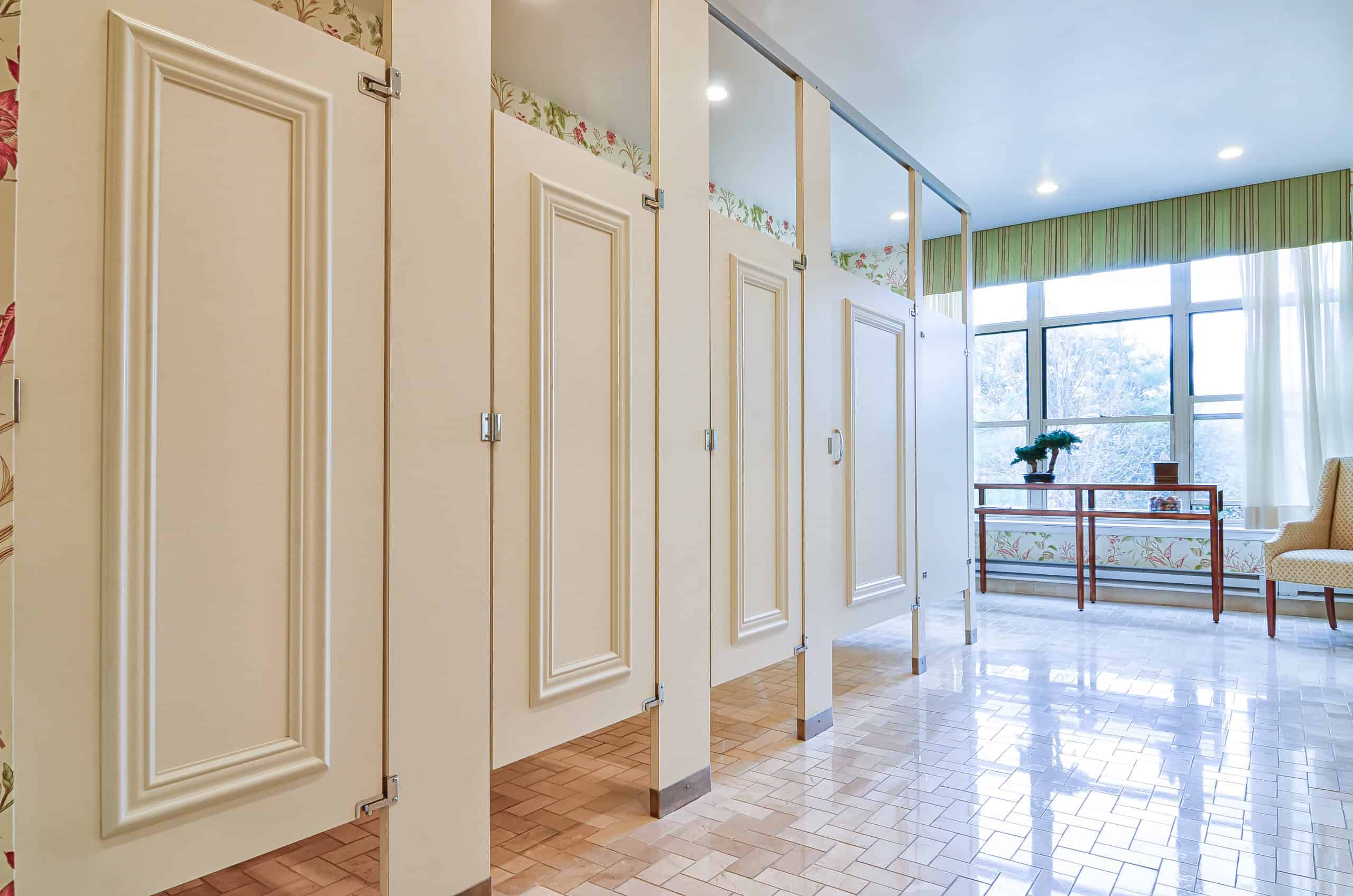 A clean public restroom with closed beige stalls, floral wallpaper, large window, striped curtains, a table, and a chair.