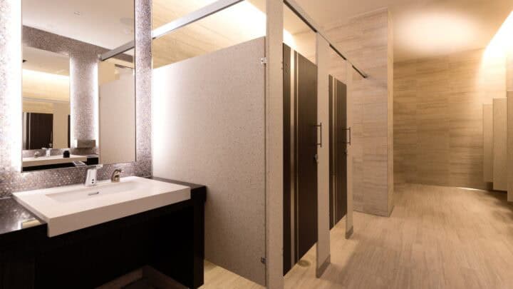 Modern public restroom with solid surface panels and woodgrain doors with metal inlays. The tiled walls are beige and there is a white sink with a large mirror on the left.