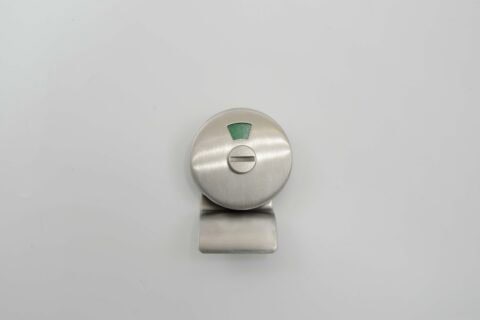 A round, brushed metal sliding lock with a green indicator on a white background, ideal for bathroom stall doors. Its design facilitates quick emergency access while maintaining a sleek appearance.
