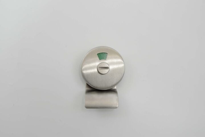A round, brushed metal sliding lock with a green indicator on a white background, ideal for bathroom stall doors. Its design facilitates quick emergency access while maintaining a sleek appearance.