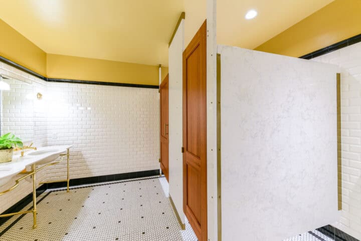 Engineered stone toilet partitions with wood veneer captured panel doors and continuous powder coated hardware, mounted floor to ceiling.