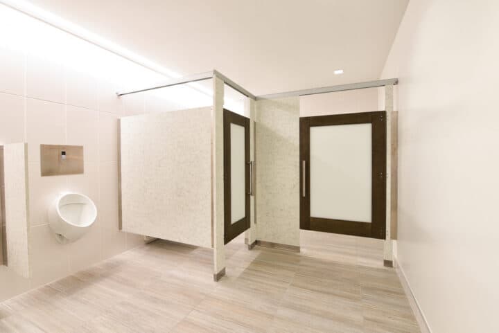 A clean, modern stone restroom with a urinal on the left and a closed stall door to the right. The room features light-colored tiles and walls, and a headrail-braced partition for added stability.
