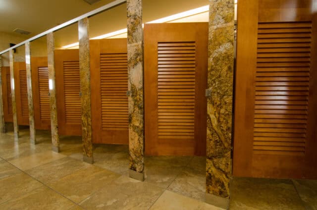A row of closed wooden bathroom stalls with louvered doors and stone dividers on a tiled floor.