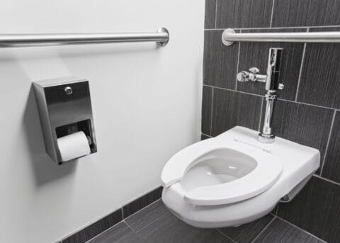 Experience the functionality of a wall-mounted toilet in a public restroom, featuring gray tiles, handrails for accessibility, and a conveniently placed toilet paper dispenser. This setup exemplifies the smallest commercial ADA bathroom layout without compromising on essential amenities.