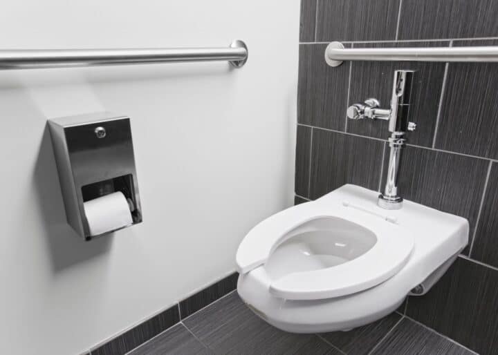 Experience the functionality of a wall-mounted toilet in a public restroom, featuring gray tiles, handrails for accessibility, and a conveniently placed toilet paper dispenser. This setup exemplifies the smallest commercial ADA bathroom layout without compromising on essential amenities.