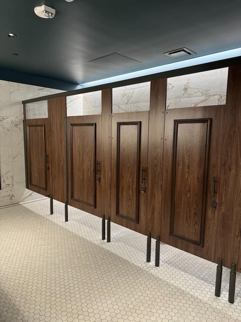 Row of wooden restroom stalls with closed doors and minimalist design, lined up against a tiled wall, with hexagonal floor tiles.