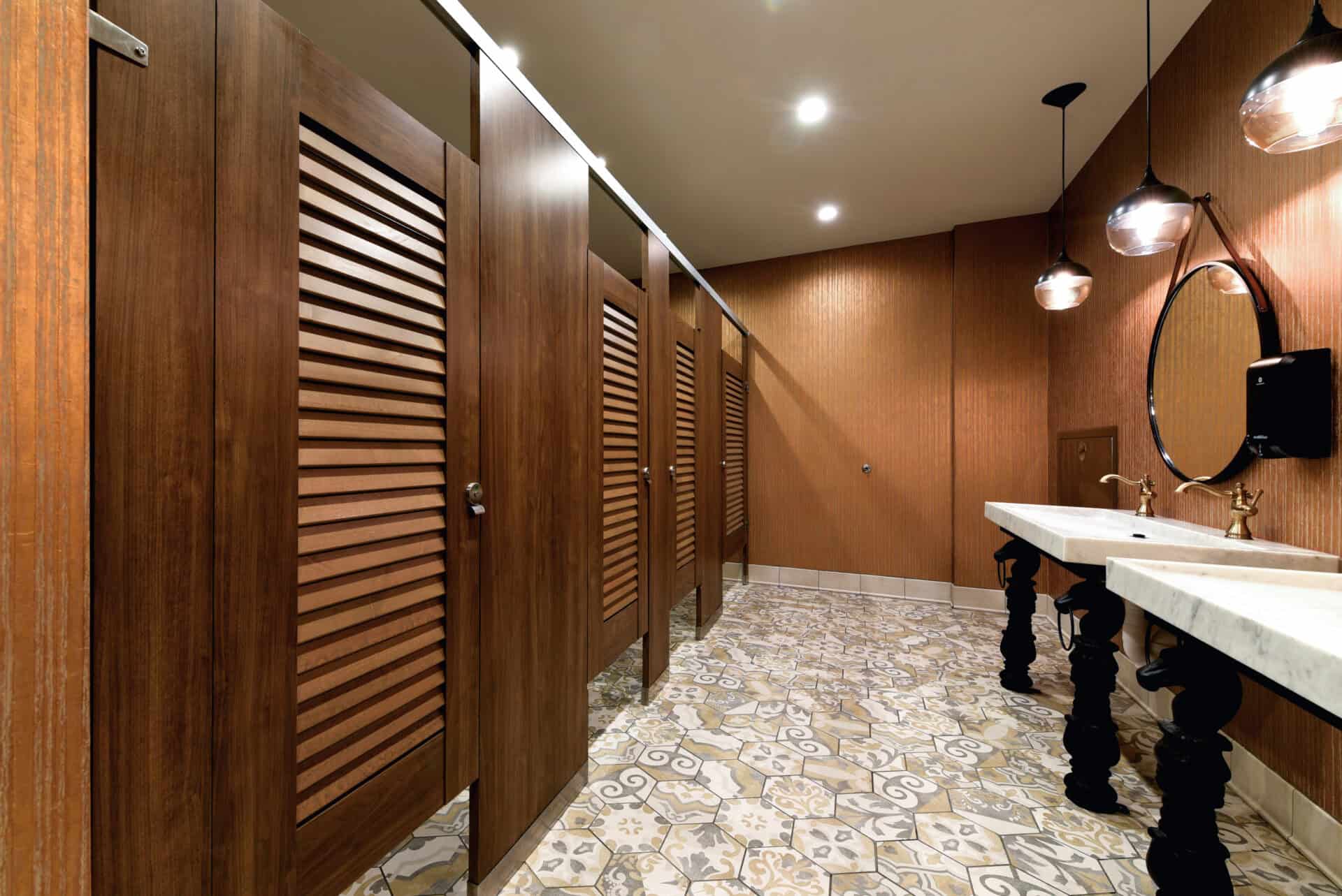 Public restroom with wooden stalls, marble sink with two basins, circular mirror, and patterned floor tiles under round ceiling lights.