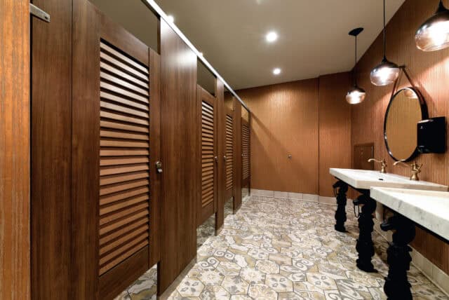 Public restroom with wooden stalls, marble sink with two basins, circular mirror, and patterned floor tiles under round ceiling lights.