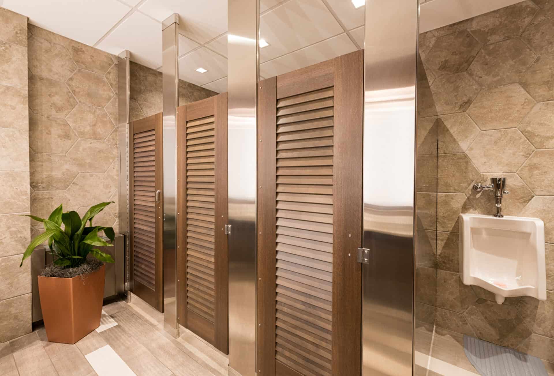 Modern men's restroom with tan tile walls, three Zero Sightline wooden stall doors for added privacy, a white urinal, and a potted green plant in the corner. A stylish Privacy Solution for commercial restroom partitions.