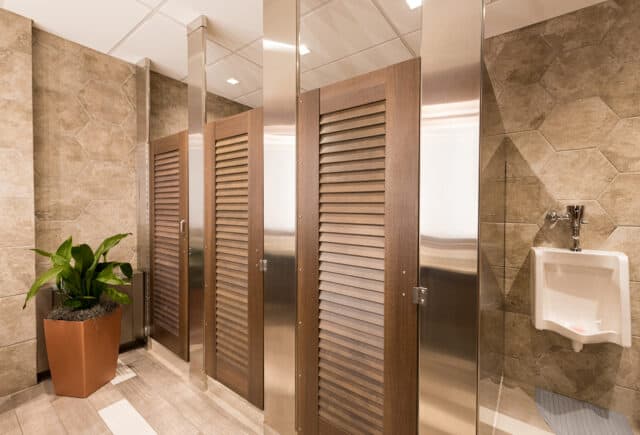 Public restroom with three closed stalls, a urinal, and a potted plant. The walls are tiled, and the floors have a wood-like appearance.