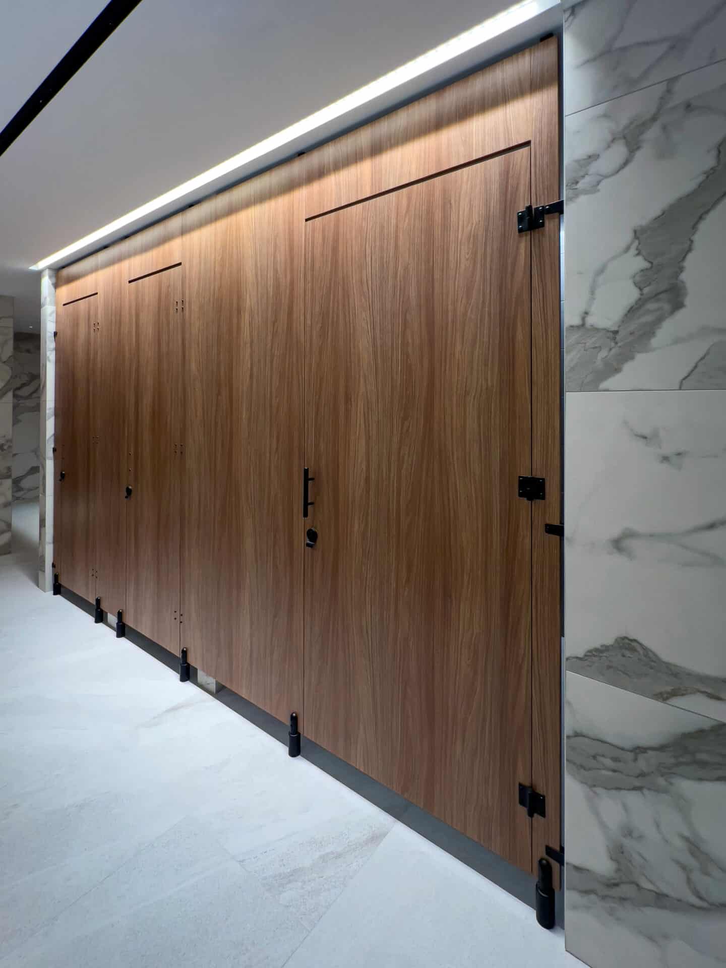 A row of modern commercial restroom partitions with wood-patterned doors and marble-patterned walls offers a sleek privacy solution, enhanced by zero sightline features and illuminated by overhead lighting.