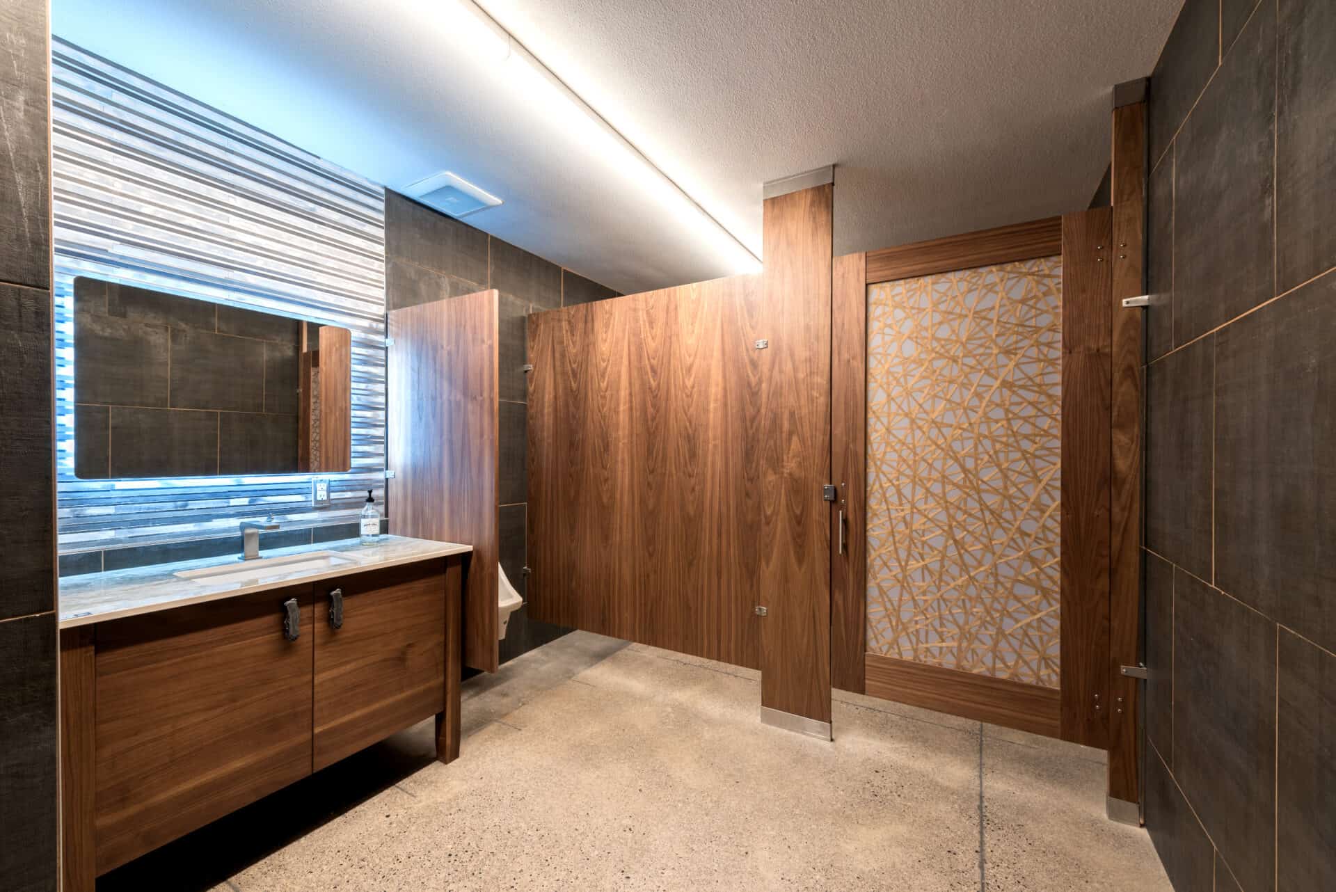 Public restroom with wooden stalls, a large mirror, and a wooden sink counter. The floor is tiled, and the lighting is bright.