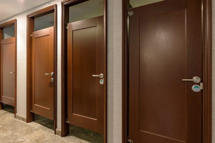 A row of four closed, brown wooden doors with silver handles in a well-lit corridor.