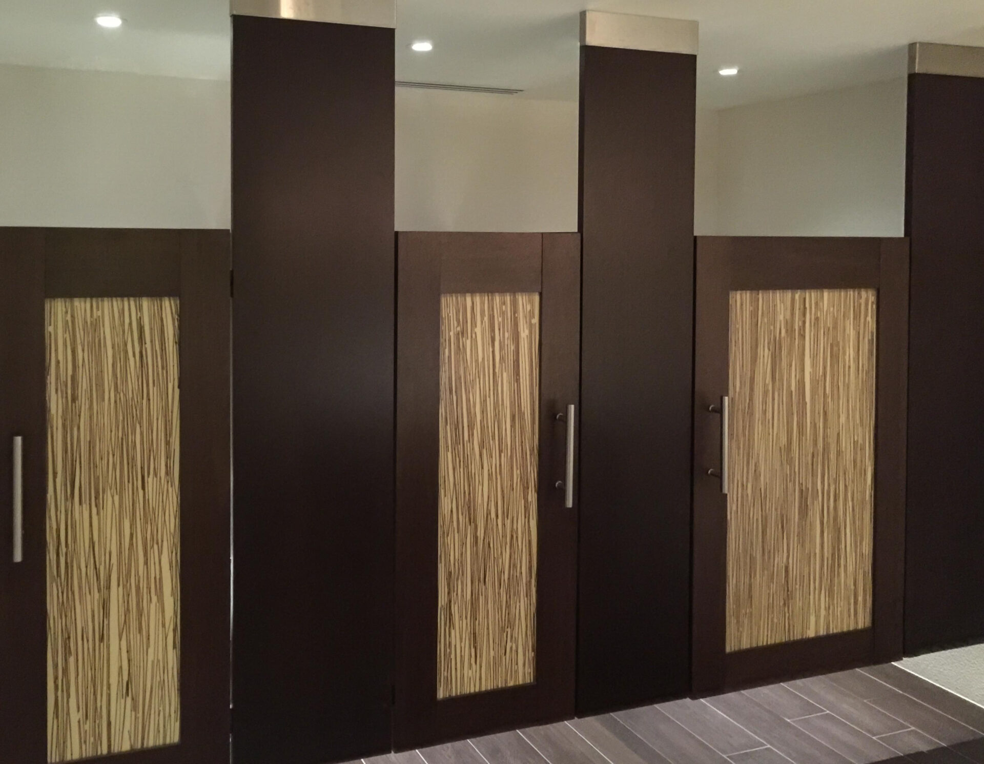 Public restroom with ceiling hung wood veneer door lite toilet partitions with embedded bamboo inserts. The floor has dark brown tiles.