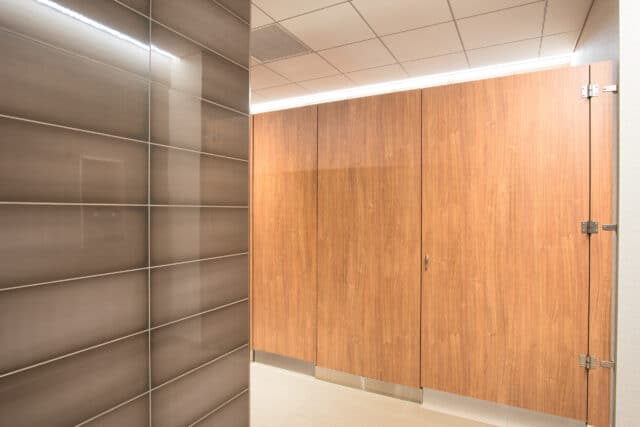 Modern public restroom featuring flat panel wood veneer floor mounted partitions and elegantly tiled walls.