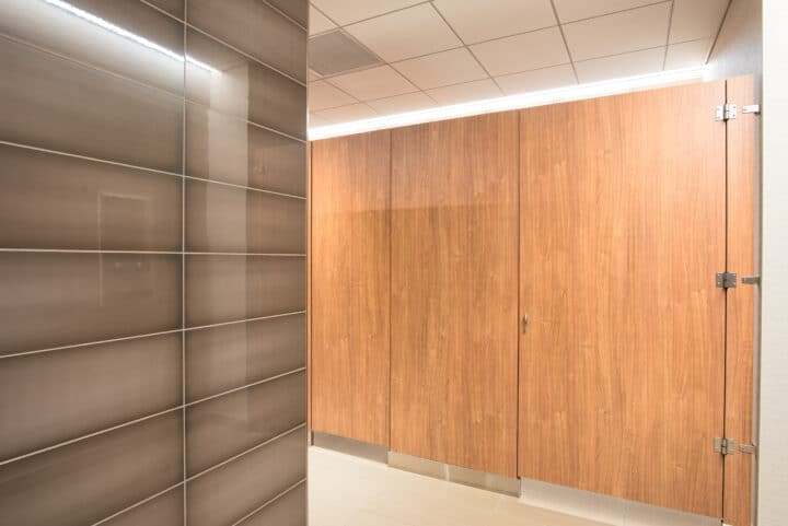 Modern public restroom featuring flat panel wood veneer floor mounted partitions and elegantly tiled walls.