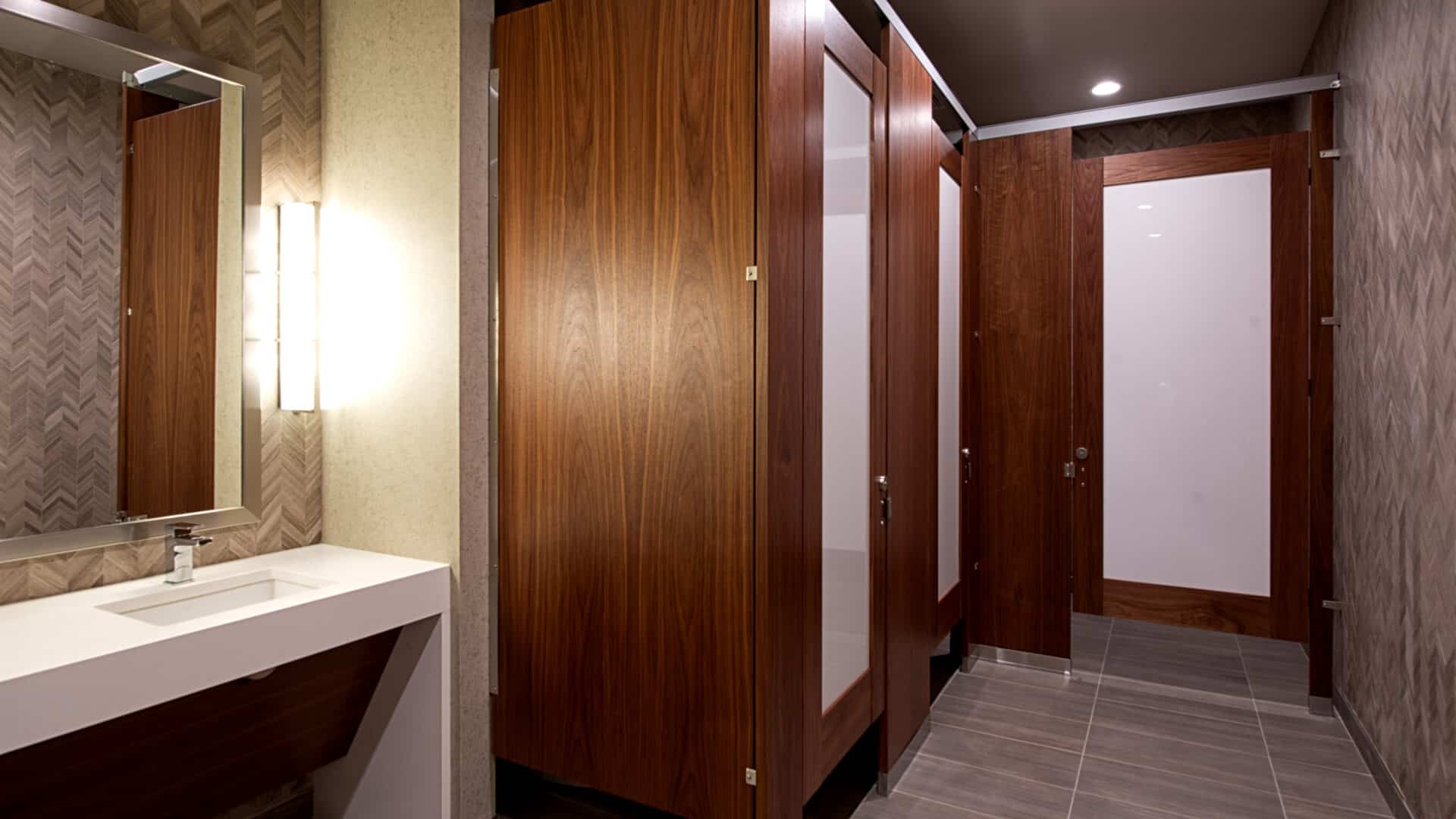 A restroom with three stalls made of wood veneer, headrail braced, with frosted acrylic door lites.