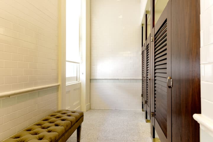 Wood veneer dressing stalls with louvered doors mounted floor to ceiling with a padded bench on the left.
