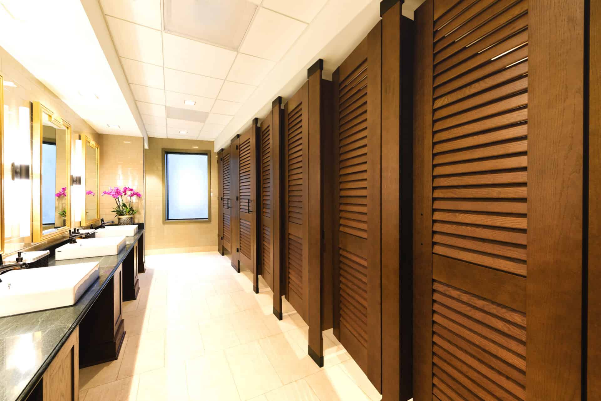University restroom with rich wood-tone louvered partitions, black hardware accents, and natural tile flooring beneath a white acoustic ceiling.