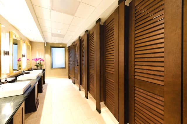 University restroom with rich wood-tone louvered partitions, black hardware accents, and natural tile flooring beneath a white acoustic ceiling.