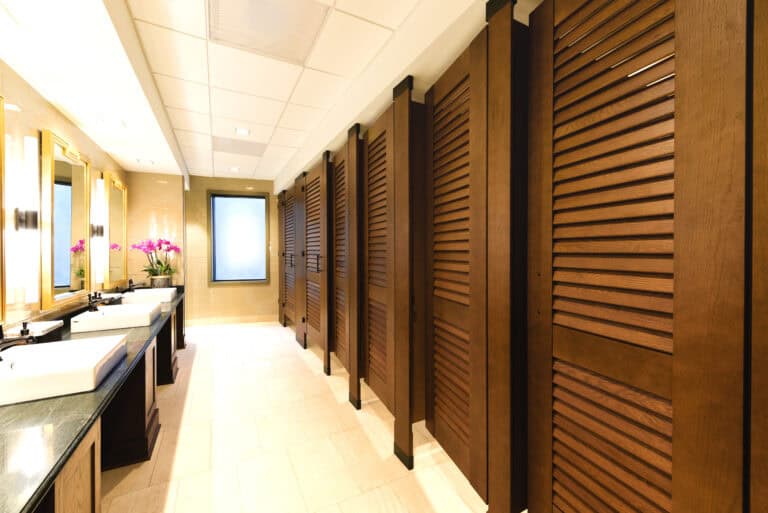 University restroom with rich wood-tone louvered partitions, black hardware accents, and natural tile flooring beneath a white acoustic ceiling.